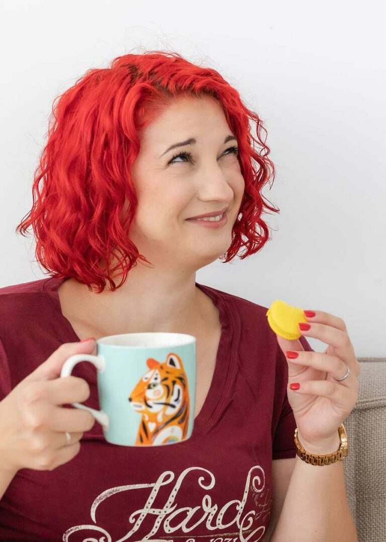 Skeptisch lächelnde Vanessa nach rechts schauend mit Tasse und Macaron in je einer Hand - Vanessa Bause Rock your Finances